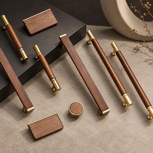 Puede incluir: Una colección de herrajes para muebles que combina madera marrón oscura y detalles en metal dorado cepillado. El conjunto incluye tiradores cilíndricos de varias longitudes, tiradores rectangulares y un pomo circular, todos con veteado.