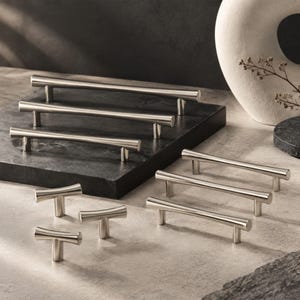 Può includere: Una collezione di maniglie per mobili in metallo color argento, che include diverse lunghe maniglie a barra e tre pomelli più piccoli a forma di T. La ferramenta presenta una finitura liscia e lucida con barre cilindriche e robusti supporti di fissaggio.
