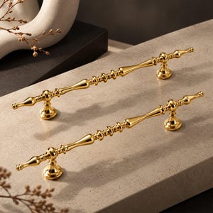 Puede incluir: Dos tiradores de armario de metal en tono dorado pulido con un diseño esbelto y alargado, detalles decorativos tipo huso y bases de montaje redondeadas. Los herrajes tienen un acabado liso y reflectante con un perfil simétrico.