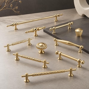 May include: A collection of gold-toned metal cabinet hardware including various drawer pulls and knobs. The set features smooth, textured, and ribbed designs with cylindrical and rounded shapes, all finished in a polished metallic gold tone.