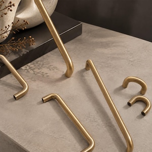 May include: A collection of brushed brass cabinet hardware including several long, slender, curved handles and two small, U-shaped pulls. Each piece features a smooth, metallic finish with rounded edges and visible screw holes at the ends.