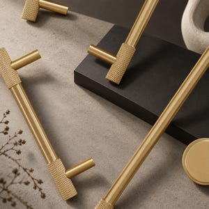 Puede incluir: Una colección de herrajes para armarios de metal dorado que incluye tiradores en forma de T y un pomo. Cada pieza presenta una barra cilíndrica lisa y un patrón moleteado texturizado en los puntos de conexión y los extremos.
