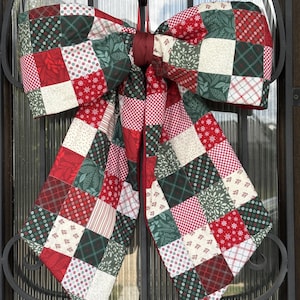 May include: A large decorative bow constructed from a patchwork of fabric squares in red, dark green, and cream. The fabric features various patterns including gingham, snowflakes, and botanical prints, all joined by a central solid red fabric knot.