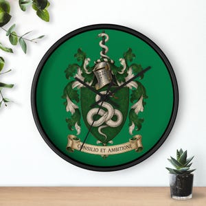 May include: A round wall clock with a black frame featuring a green background, a central shield with a white snake, a silver knight's helmet, and a banner at the bottom with the Latin text "CONSILIO ET AMBITIONE" in black capital letters.