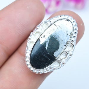 Puede incluir: Un anillo de forma ovalada con una piedra pulida de dos tonos, negro y plata metálica, engastada en una banda de plata decorativa. El engaste tiene un borde con cuentas y una serie de pequeños recortes circulares alrededor.