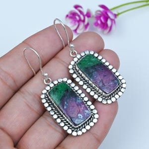 May include: A pair of drop earrings featuring rectangular stones with a mix of green and purple hues. Each stone is set in a silver-toned metal frame with a beaded border and a twisted rope detail, suspended from simple silver-toned ear wires.