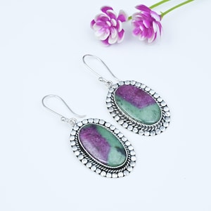 Può includere: Un paio di orecchini pendenti di forma ovale con pietre preziose di rubino zoisite dai vivaci toni magenta e verde. Ogni pietra è incastonata in una montatura in metallo argentato con un bordo decorativo a perline e un gancio curvo.