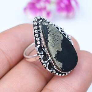 Puede incluir: Un anillo en forma de lágrima con una piedra de pirita metálica oscura engastada en una banda de metal plateado. El engaste está decorado con un borde texturizado y con cuentas que enmarca la piedra, creando un borde detallado alrededor.
