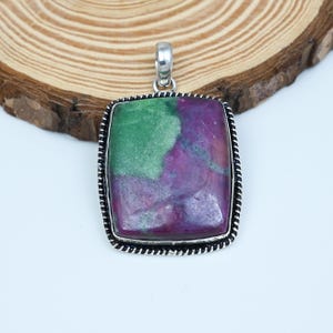 Puede incluir: Un colgante rectangular con una piedra pulida que presenta secciones distintas en verde y magenta intenso. La piedra está engastada en un marco de metal plateado con un borde de estilo cuerda trenzada y una anilla plateada superior.