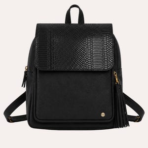 May include: A black backpack featuring a textured snake-skin pattern on the top flap and a smooth finish on the main body. The bag includes a gold-toned zipper pull with a long black tassel, adjustable shoulder straps, and a small gold logo emblem.