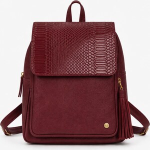 May include: A deep burgundy backpack featuring a textured snakeskin-patterned flap closure. The bag includes a front pocket, a gold-toned circular logo emblem, and a matching burgundy tassel attached to the side zipper pull with gold hardware.