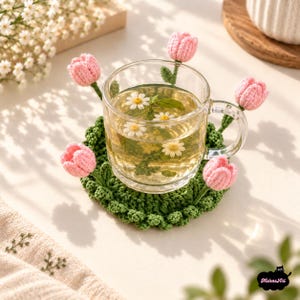 Puede incluir: Una taza de cristal transparente llena de té y pequeñas flores de margarita blancas sobre un posavasos redondo de ganchillo verde oscuro. El posavasos presenta cuatro pequeños capullos de tulipán de ganchillo rosa claro en los bordes.