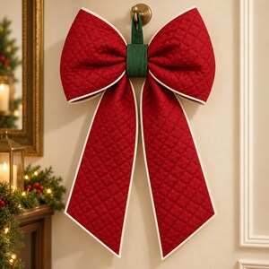 May include: A large, quilted red fabric bow with white piping edges and a dark green center loop. The bow features a diamond-stitched pattern throughout the fabric and is suspended from a small, circular gold-toned metal wall hook.