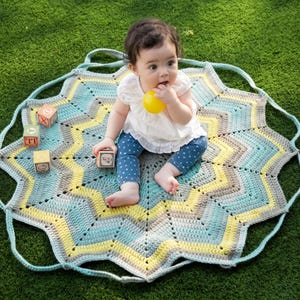 May include: A star-shaped crochet play mat featuring concentric rings of light blue, yellow, and grey yarn. The mat has integrated handles and is decorated with three wooden alphabet blocks and a small yellow ball.