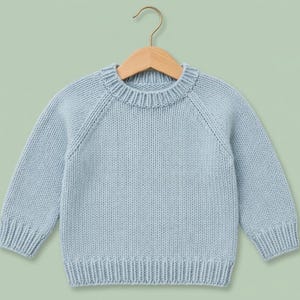 Pode incluir: Uma camisola de malha de manga comprida em azul claro com gola redonda canelada, punhos canelados e bainha canelada. A camisola é feita com mangas raglan e está exposta num cabide de madeira, apresentando um design simples e liso.