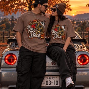 May include: A tan t-shirt featuring a graphic print of a car wheel, a ghost, and pumpkins with the text The Nice Stock. The design includes autumn leaves and bats. The shirt is worn with black cargo pants and white sneakers in a fall setting.