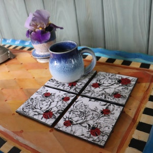 May include: Set of four black and white coasters with a floral design and red ladybugs. The coasters are on a wooden table with a blue and white checkered pattern.