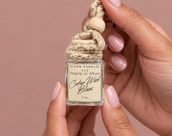 May include: A small square glass car diffuser bottle with a beige label reading Olson Candles LLC, Hanging car diffuser, Cedar Wood Blanc, and 7 ml. The bottle features a braided beige rope with gold metallic threading and a wooden bead topper.