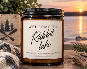 May include: An amber glass jar candle with a black lid and a white label featuring the text "WELCOME TO Rabbit lake" in black, along with "Olson Candles LLC | 227g soy wax | Caribbean Teakwood" and the website address printed at the bottom.