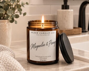 May include: A lit soy wax candle in an amber glass jar with a black lid. The white label reads OLSON CANDLES LLC, www.olsoncandles.com, Magnolia & Peony, Hand poured soy wax candle, 100% natural, 227g | 8oz. The candle has a single burning wick.