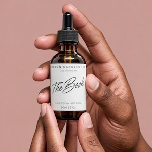 May include: A 2 oz amber glass dropper bottle with a black cap and a white label. The label reads OLSON CANDLES LLC, Des Moines, IA, The Book, and Card diffuser refill bottle, 60ml | 2 Oz. The bottle contains a liquid and has a dropper top.