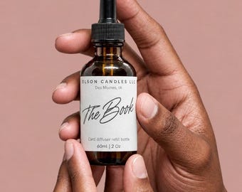 May include: A 60ml amber glass dropper bottle with a black cap and a white label. The label reads OLSON CANDLES LLC, Des Moines, IA, The Book, and Card diffuser refill bottle, 60ml | 2 Oz. The bottle contains a liquid and has a dropper top.