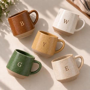 Puede incluir: Cinco tazas de cerámica con acabado moteado y base acanalada. Cada taza presenta una letra en relieve: B en una taza marrón, W en una blanca, Y en una amarilla, G en una verde y E en una beige claro. Cada una tiene un asa lateral.