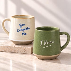 Op de afbeelding: Twee keramische mokken met oren. De crèmekleurige mok heeft de blauwe tekst "You Complete Me". De groene mok heeft de witte tekst "I Know" met een onderstreping. Beide mokken hebben een gespikkelde afwerking en een ongeglazuurde onderrand.