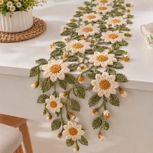 May include: A long, rectangular crochet table runner featuring a repeating pattern of white daisy flowers with yellow centers, connected by intricate green crocheted leaves and small, closed flower buds along the entire length of the piece.