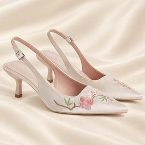 May include: A pair of cream-colored satin slingback heels with a pointed toe and a low kitten heel. The shoes feature delicate floral embroidery in shades of pink and green on the front, with an adjustable buckle strap at the heel.