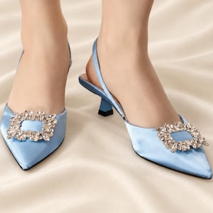 May include: Light blue satin slingback pumps with a pointed toe and a low kitten heel. Each shoe features a large, rectangular decorative buckle on the front, encrusted with clear, multifaceted crystals that catch the light.