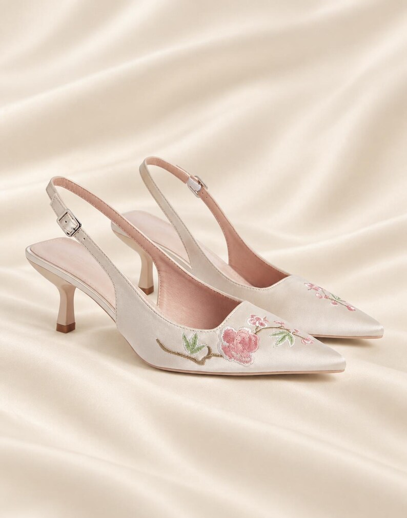 May include: A pair of cream-colored satin slingback heels with a pointed toe and a low kitten heel. The shoes feature delicate floral embroidery in shades of pink and green on the front, with an adjustable buckle strap at the heel.