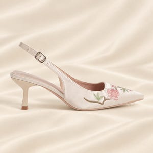 May include: A cream-colored slingback pump with a pointed toe and a low kitten heel. The shoe features delicate floral embroidery in shades of pink and green on the toe area, an adjustable ankle strap with a silver buckle, and a smooth finish.