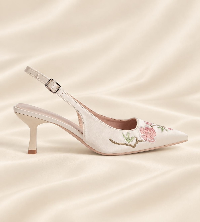 May include: A cream-colored slingback pump with a pointed toe and a low kitten heel. The shoe features delicate floral embroidery in shades of pink and green on the toe area, an adjustable ankle strap with a silver buckle, and a smooth finish.