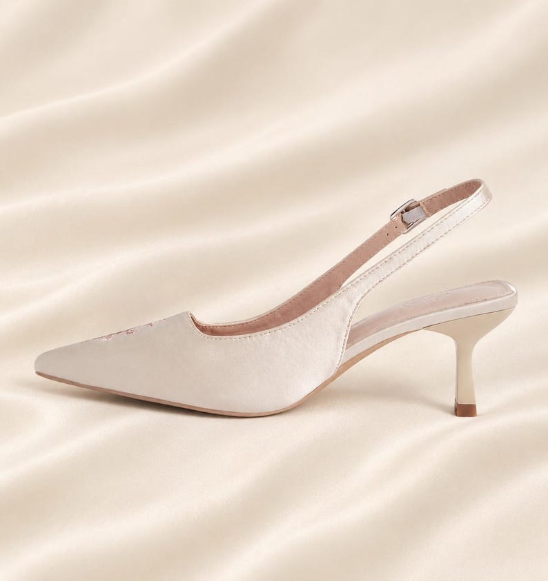 May include: A champagne-colored satin slingback pump with a pointed toe and a low kitten heel. The shoe features a thin adjustable ankle strap with a small silver-toned metal buckle and a smooth, light-colored interior lining.