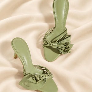 May include: A pair of sage green heeled sandals featuring a slip-on design with a kitten heel. The front strap is adorned with multiple thin cord-like straps gathered into a decorative knot detail with small knotted ends and leaf-shaped accents.