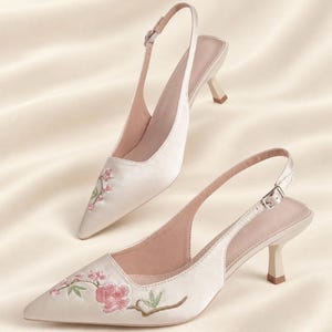 May include: A pair of cream-colored satin slingback heels with a pointed toe and a kitten heel. The shoes feature delicate pink floral embroidery with green leaves on the front. Each shoe has an adjustable ankle strap with a silver buckle.