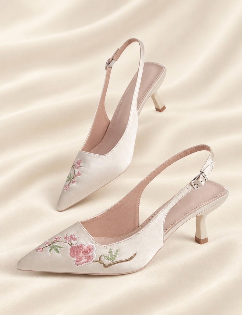 May include: A pair of cream-colored satin slingback heels with a pointed toe and a kitten heel. The shoes feature delicate pink floral embroidery with green leaves on the front. Each shoe has an adjustable ankle strap with a silver buckle.
