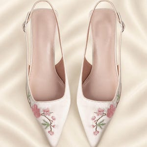 May include: A pair of pale pink pointed-toe slingback shoes featuring delicate floral embroidery with pink blossoms and green stems on the toe area. The shoes have a slim adjustable ankle strap with a small silver-toned metal buckle on each side.