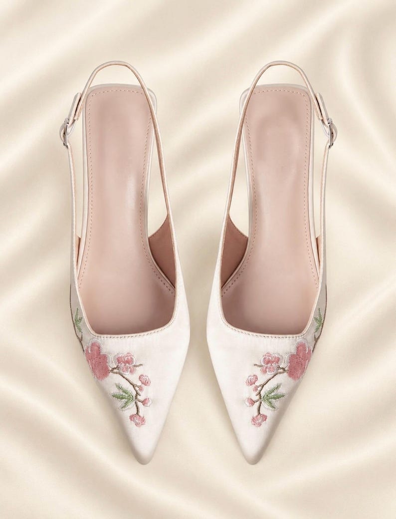 May include: A pair of pale pink pointed-toe slingback shoes featuring delicate floral embroidery with pink blossoms and green stems on the toe area. The shoes have a slim adjustable ankle strap with a small silver-toned metal buckle on each side.