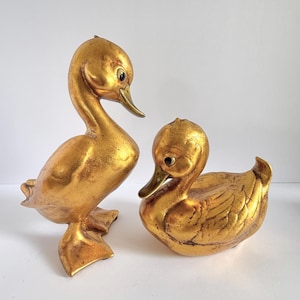May include: Two gold-colored duck figurines with a distressed metallic finish. One duck is standing upright with its head turned, while the other duck is in a seated position. Both figures feature dark eyes and smooth, curved body shapes.