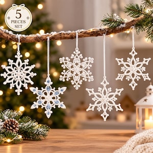 Puede incluir: Un conjunto de cinco adornos de copos de nieve blancos colgados de una rama de madera. Cuatro adornos son de color blanco sólido, mientras que uno presenta detalles en azul claro. Cada copo tiene patrones de encaje y un cordón blanco.