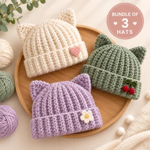 May include: A bundle of three crocheted hats with cat ears. One hat is cream with a pink heart, one is sage green with two red cherries, and one is lavender with a white flower. Each hat features a ribbed texture and a folded brim design.