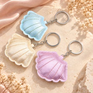 May include: Three seashell-shaped keychains in metallic light blue, gold, and light pink. Each keychain features a stitched shell design, a silver-toned metal jump ring, and a circular split ring for keys, crafted from a smooth, reflective material.