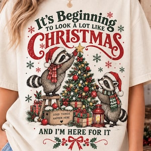 May include: A cream-colored t-shirt featuring a graphic of two raccoons decorating a Christmas tree. The text reads, "It's Beginning to look a lot like CHRISTMAS AND I'M HERE FOR IT." The design includes presents, a lantern, and snowflakes.