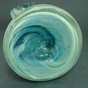 May include: A circular glass vase base featuring a swirling pattern of translucent teal, light blue, and white colors. The glass has a smooth, polished surface with visible internal air bubbles and a central pontil mark on the bottom.