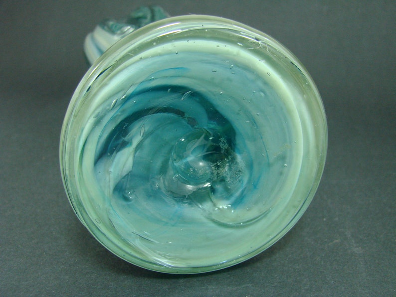 May include: A circular glass vase base featuring a swirling pattern of translucent teal, light blue, and white colors. The glass has a smooth, polished surface with visible internal air bubbles and a central pontil mark on the bottom.