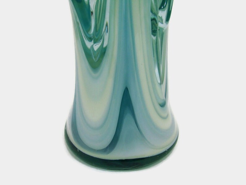 May include: A glass vase featuring a swirling pattern of teal, light blue, and white colors. The surface has a smooth, glossy finish with vertical, elongated streaks that create a fluid, marbled appearance throughout the cylindrical body.