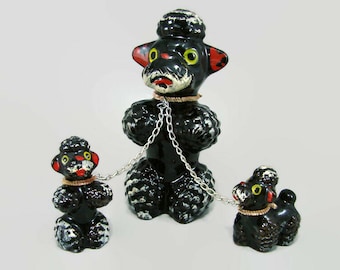 May include: A set of three black ceramic poodle figurines with white painted accents on their fur. The larger central poodle is connected by a silver metal chain to two smaller poodle figurines, one on each side, each wearing a tan collar.