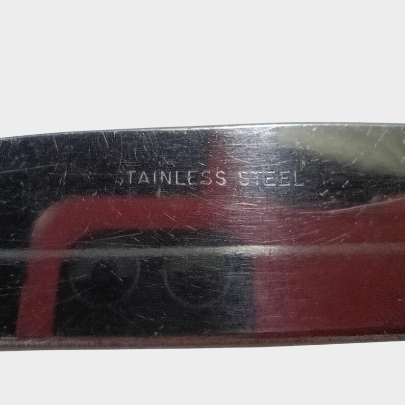 Puede incluir: Primer plano de una hoja de cuchillo de acero inoxidable. El texto "STAINLESS STEEL" est&aacute; grabado en el metal. La hoja tiene una superficie reflectante con reflejos rojos y negros.